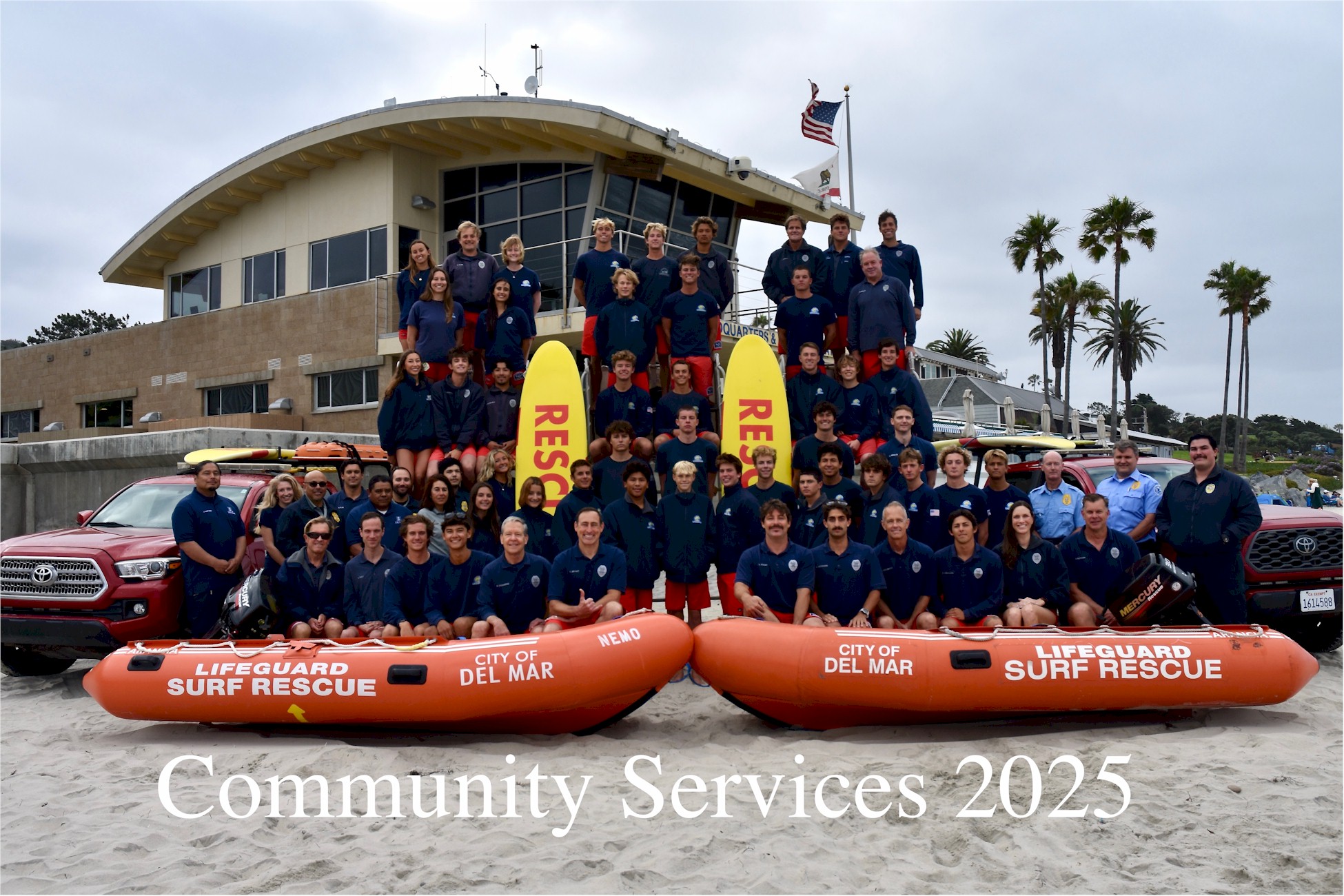 2025 Del Mar lifeguard team photographed on the sand in front of lifeguard headquarters with two rescue boats.